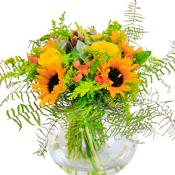 The bouquet at the centre of this image speaks immediately of sunlit abundance, its vivid palette dominated by plush, yellow-orange sunflowers and garden roses that glow with the exuberance of midsummer afternoons in Queen's Park. Dense, radiant petals unfurl in layers, each one meeting the next in a celebration of natural artistry. Weaving through the arrangement, jewel-like Hypericum berries in deep red provide unexpected bursts of colour, their gloss a lively counterpoint to the soft, inviting petals of roses and sunflowers. A tapestry of foliage supports and elevates the blooms: delicate fern fronds curve outward and downward in lively sweeps, displaying the kind of movement seen in the tall grasses near Queen's Park station. Sprigs of light green filler-perhaps goldenrod dotted with its tiniest blooms-and wispy, silver-blue eucalyptus leaves add airiness and contrast, blurring the boundaries between meadow and modern design. The elegantly rounded glass vase cradles every stem, submerged in clear water that seems to capture and magnify the arrangement's vitality. Up close, you'd sense the velvety touch of petals, the fresh, grassy aroma mingling with hints of woodland trails nearby. Designed for local celebrations-be it a milestone, birthday, or just because-this bouquet reflects Queen's Park's sense of community and the joy that nature's beauty brings to every gathering.